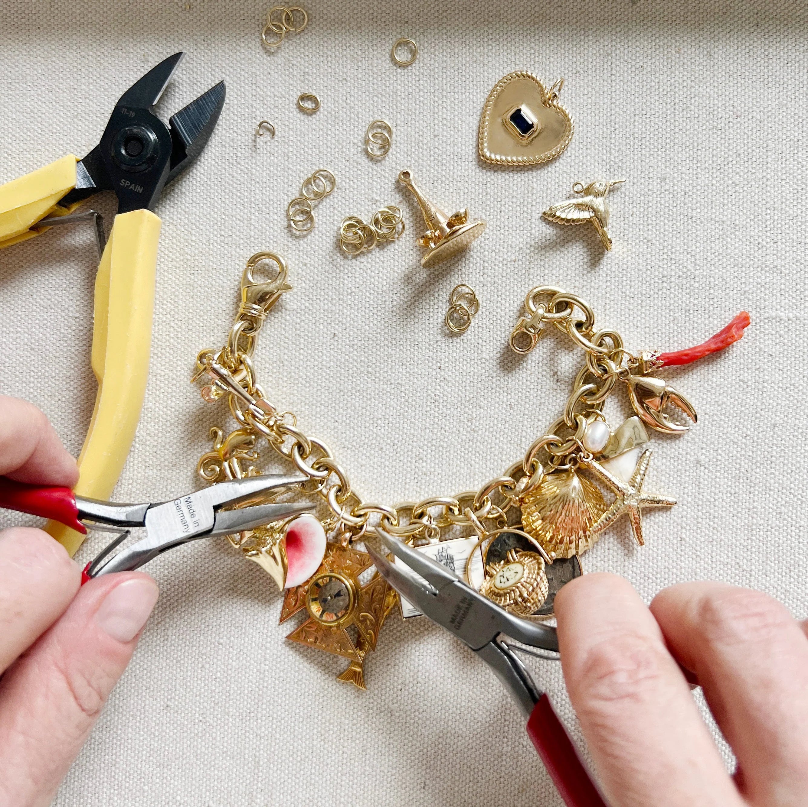 Gold charm bracelet being worked on with pliers and various charms on a light background
